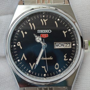 Pode incluir: Um relógio de pulso automático Seiko 5 em aço inoxidável prateado com mostrador azul escuro com numerais árabes orientais. O relógio inclui uma janela de dia e data na posição das três horas exibindo MON 23 e uma pulseira de elos de metal prateado.