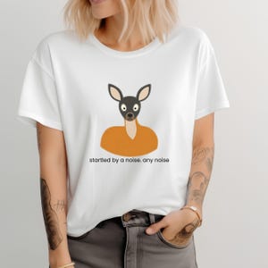 May include: A white short-sleeve t-shirt featuring a graphic of a deer head with large eyes and a tan body. Below the graphic, the text reads: startled by a noise. any noise. The shirt has a crew neckline and a relaxed fit, made of cotton fabric.