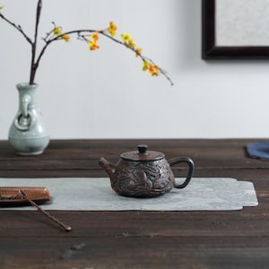 May include: A dark brown clay teapot featuring intricate relief carvings of a dragon and phoenix. The teapot has a rounded body, a curved spout, a black handle, and a matching lid with a small knob. It sits on a light gray rectangular mat.