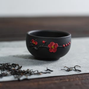 May include: A small, black ceramic tea cup with a floral design. The cup features a raised relief of red flowers and buds on a branch. The cup is round and sits on a light gray surface.