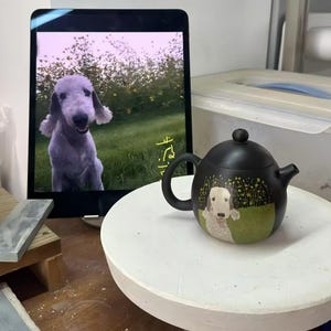 May include: A black ceramic teapot with a rounded body, a curved handle, and a spout. The teapot features a painted illustration of a dog in a field of flowers. The teapot is sitting on a white surface.