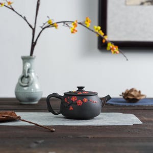 May include: A black ceramic teapot with a curved handle and spout, decorated with red and yellow floral designs. The teapot sits on a gray surface, with a vase and decorative branch in the background. The teapot is a traditional style.