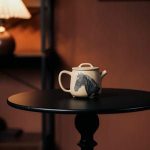 May include: A beige ceramic teapot featuring a black horse head design. The teapot has a curved handle, a small spout, and a lid with a loop handle. It sits on a black round table.
