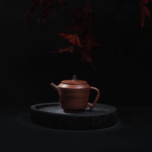 May include: A brown ceramic teapot with a lid and a small handle, set on a dark wooden tray. The teapot has a spout and a curved handle. The background is dark with red leaves.