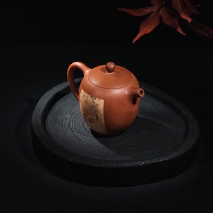 Puede incluir: Una tetera de cerámica de color marrón rojizo con un cuerpo redondeado, un asa curva y un pequeño pico. La tetera tiene una tapa con un pequeño pomo y una etiqueta decorativa. Se encuentra sobre una bandeja de madera oscura y redonda, con un fondo oscuro y una hoja roja.