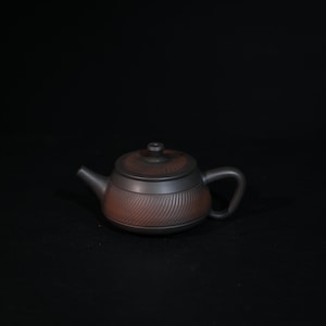 May include: A dark brown clay teapot featuring a rounded body and a matching lid. The exterior surface is decorated with a fine, diagonal ribbed texture that wraps around the midsection of the pot and the top of the lid.