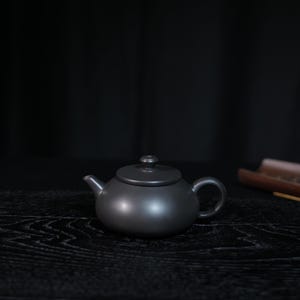 May include: A matte black ceramic teapot with a rounded body, a short curved spout, a circular lid with a small knob handle, and a C-shaped handle. The teapot has a smooth, uniform finish and a low-profile design, presented in a simple form.