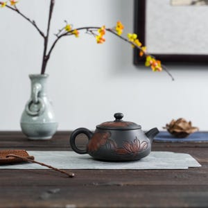 Könnte beinhalten: Eine schwarze Keramik-Teekanne mit einem rotbraunen Blumendesign. Die Teekanne hat einen gebogenen Griff und Ausguss sowie einen passenden Deckel. Im Hintergrund befindet sich eine Vase mit einem Zweig aus gelben und orangen Blüten.
