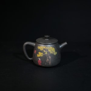 May include: A dark gray, textured ceramic teapot with a matching lid, a curved handle, and a short spout. The body features a painted illustration of a yellow pine tree and a small red figure, along with a red square stamp mark on the side.