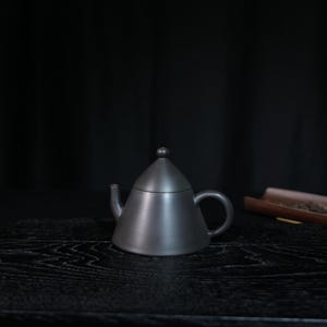 May include: A matte black teapot with a conical body and a matching lid featuring a small spherical knob. The teapot has a curved handle on the right side and a short, straight spout on the left, presenting a smooth, uniform dark finish throughout.