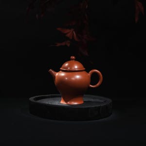 Puede incluir: Una tetera de cerámica de color marrón rojizo con cuerpo redondeado, boquilla y asa. La tetera tiene una tapa a juego con un pequeño pomo en la parte superior. Se encuentra sobre una bandeja de madera redonda y oscura sobre un fondo oscuro.