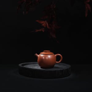 May include: A small, reddish-brown teapot with a lid and a curved handle, presented on a dark, circular wooden tray. The background is a dark, blurred scene with hints of red leaves. The teapot is likely made of ceramic.