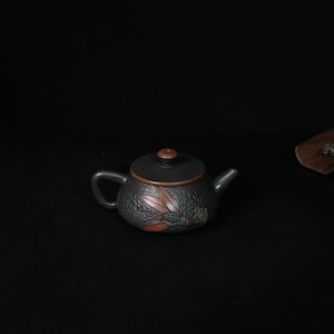 May include: A dark brown clay teapot with a textured, pebbled surface. The teapot features a smooth, flat lid with a small knob and a decorative relief of reddish-brown leaves on the side. It has a curved handle and a short, tapered spout.