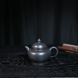 May include: A matte black teapot with a rounded body, a curved handle, a short spout, and a matching lid featuring a small, pointed knob. The teapot has a smooth, uniform finish and is centered in the frame against a dark background.