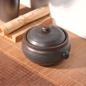 May include: A dark brown ceramic lidded jar with a rounded shape and a small knob handle on the lid. The exterior features horizontal bands of textured, wavy line patterns and a small, sculpted animal head detail on the side of the vessel.