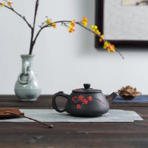 Könnte beinhalten: Schwarze Keramik-Teekanne mit roten Blumenmustern. Die Teekanne hat einen gebogenen Griff und Ausguss und steht auf einer grauen Oberfläche. Im Hintergrund befindet sich eine Vase mit einem Zweig gelber Blumen.