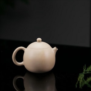 May include: A round, light beige teapot with a smooth, matte finish. The teapot features a curved handle, a short spout, and a matching lid topped with a small, spherical knob. The design is simple and spherical, showcasing a uniform, neutral color.