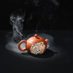 Könnte beinhalten: Terrakottafarbene Teekanne mit Tiger-Illustration. Die Teekanne hat einen runden Körper, einen gebogenen Griff und einen kleinen Ausguss. Dampf steigt aus der Teekanne vor dunklem Hintergrund auf.