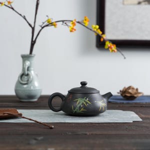 May include: A black ceramic teapot with a lid, handle, and spout. The teapot is decorated with green bamboo leaves and stems. A small vase with a branch of yellow flowers is in the background.