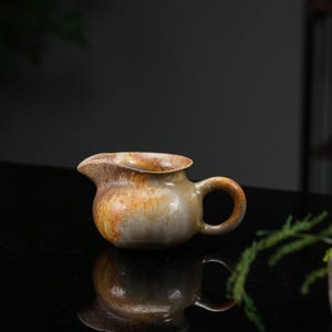 Puede incluir: Una jarra de cerámica para leche con cuerpo redondeado, un pequeño pico vertedor y un asa curva. El exterior presenta un esmalte moteado con tonos tierra de marrón, beige y gris, creando una apariencia texturizada en la superficie.