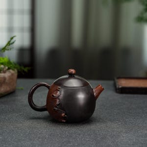May include: A dark brown teapot with a smooth, rounded body and a matching lid featuring a small knob. The side of the teapot displays a textured, leaf-like relief pattern in a reddish-brown tone, complemented by a curved handle and a short spout.