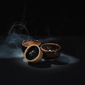 May include: Three small, round ceramic bowls with a dark, mottled brown glaze and a smooth, glossy interior finish. The bowls are arranged in a cluster, surrounded by wisps of white smoke that curl around the exterior surfaces against a dark background.