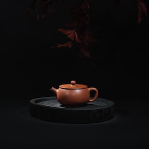 Op de afbeelding: Een kleine, roodbruine keramische theepot met een deksel en handvat staat op een donker, rond houten dienblad. De theepot heeft een tuit en een kleine knop op het deksel. De achtergrond is donker en wazig.