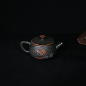 May include: A dark charcoal gray teapot with a textured surface and a smooth reddish-brown lid. The body features a relief design of reddish-brown leaves and a small round fruit. It has a curved handle and a short, tapered spout.