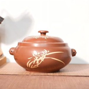 May include: A round, reddish-brown ceramic pot with a matching lid and two small side handles. The exterior features a light-colored, painted floral design with long, slender leaves and blossoms on the front and top of the lid.