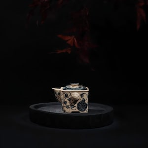 Peut inclure: Une théière carrée en céramique avec une base de couleur crème ornée de motifs floraux et de vignes bleu foncé. La théière comprend un couvercle assorti avec une bordure géométrique, un petit bec verseur latéral et une attache de poignée.