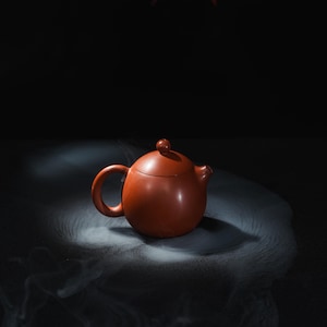 Puede incluir: Una pequeña tetera redonda, de color marrón rojizo, con tapa a juego y asa curvada. La tetera tiene un pequeño pico y un pomo en la parte superior de la tapa. El fondo es oscuro, con un efecto suave y ahumado alrededor de la tetera.
