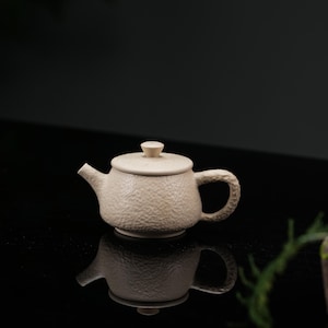 May include: A beige ceramic teapot with a textured, hammered-style surface. The teapot features a rounded body, a short spout, a curved handle with a matching hammered finish, and a circular lid topped with a small, knob-like handle.