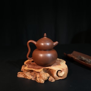 Op de afbeelding: Een bruine kleien theepot in de vorm van een dubbele kalebas, voorzien van een gebogen handvat, een kleine tuit en een deksel met een knop. De theepot heeft een gladde, matte afwerking en staat op een natuurlijk, onregelmatig stuk hout.