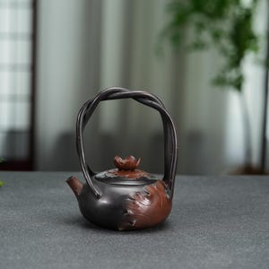 Puede incluir: Una tetera de cerámica de color marrón oscuro y negro con un asa alta y arqueada que recuerda a ramas retorcidas. La tapa está rematada con un pequeño capullo de flor esculpido y el cuerpo de la tetera está decorado con patrones de hojas en relieve.