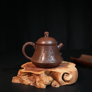 Könnte beinhalten: Eine braune Tonteekanne mit einem strukturierten Griff, einem Deckel mit kleinem skulpturalem Knauf und einem eingravierten Motiv auf dem Körper, das einen Schmetterling und Bambusstängel zeigt. Die Teekanne hat einen breiten Boden und einen kurzen Ausguss.