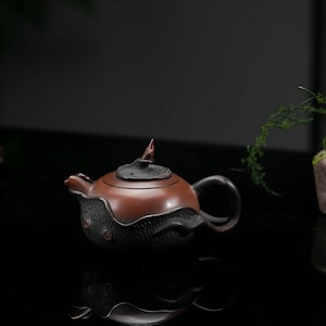 May include: A two-toned teapot featuring a smooth, reddish-brown upper body and a textured, dark charcoal-colored lower section. The lid is topped with a small, pointed finial, and the handle is curved, matching the dark, textured base.