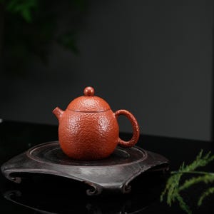 May include: A round, reddish-brown teapot with a textured, dimpled surface. The teapot features a small, spherical lid knob, a curved spout, and a rounded handle. The exterior has a consistent, granular finish across the body, lid, and handle.