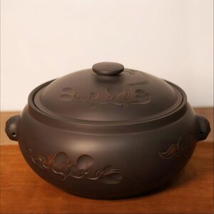 May include: A dark brown ceramic pot with a matching lid, featuring carved floral vine patterns on the exterior surface. The pot has two small side handles and a rounded body shape, with a smooth matte finish across the entire exterior.