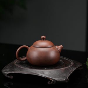 Peut inclure: Une théière en argile brun foncé de forme ronde avec une finition mate et lisse. Elle présente une anse incurvée, un bec court et un couvercle assorti surmonté d'un petit bouton arrondi. Le corps est bombé et repose sur un support en bois sombre.
