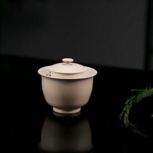 Puede incluir: Un cuenco de té gaiwan de cerámica en color blanco roto mate con tapa a juego. El cuenco presenta un borde ancho y acampanado y una base circular pequeña. La tapa tiene un pequeño pomo central. El diseño es sencillo, liso y uniforme.