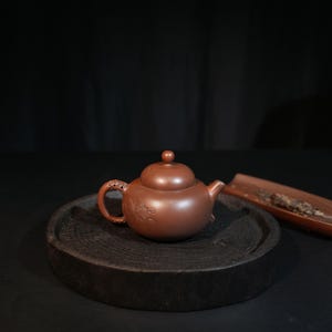 May include: A round, reddish-brown clay teapot featuring a small knob on the lid, a curved spout, and a handle with intricate relief carvings of a fish. The teapot sits on a dark, circular wooden base with a visible grain texture.