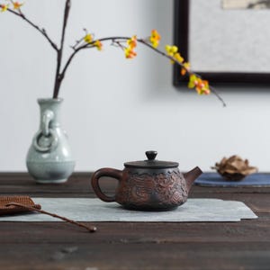 以下が含まれることがあります： 茶色のセラミックティーポット。濃い色の蓋とハンドルが付いています。 ティーポットはエンボス加工のデザインで、灰色のマットの上に置かれています。 黄色の花の枝が入った花瓶が背景にあります。 ティーポットは装飾品です。