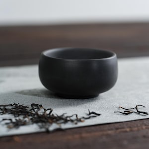 Puede incluir: Una pequeña taza de té de cerámica negra mate se encuentra sobre una superficie gris claro, con hojas de té secas esparcidas alrededor. La taza tiene una forma redondeada y una superficie lisa y sin adornos, lo que sugiere un diseño minimalista.