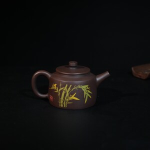 May include: A dark brown clay teapot featuring a cylindrical body, a curved handle, a short spout, and a flat lid with a small knob. The side of the teapot is decorated with a yellow and green bamboo stalk illustration and a small red square seal.