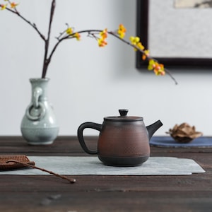 Op de afbeelding: Een bruine en zwarte keramische theepot met een textuuroppervlak, een gebogen handvat en een tuit. De theepot staat op een grijze mat op een donkere houten tafel. Op de achtergrond een vaas met een tak met gele bloemen.