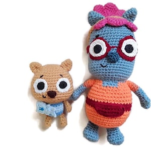 May include: Two handmade crochet dolls. One is a light brown bear-like creature with a blue bow tie. The other is a blue creature with red-rimmed glasses, an orange top, and a pink headpiece. Both dolls have black and white eyes.