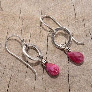 Peut inclure: Une paire de boucles d'oreilles en argent avec une texture martelée. Chaque boucle d'oreille présente un petit anneau rond en argent avec un pendentif en pierre précieuse de couleur rubis enroulé de fil. Les boucles d'oreilles sont exposées sur une surface en bois.