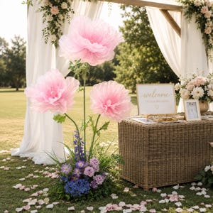 May include: Three large, light pink artificial flowers on tall green stems, arranged with a base of purple and blue flowers and greenery. Next to them is a rectangular wicker table holding a white sign that reads welcome to our wedding.