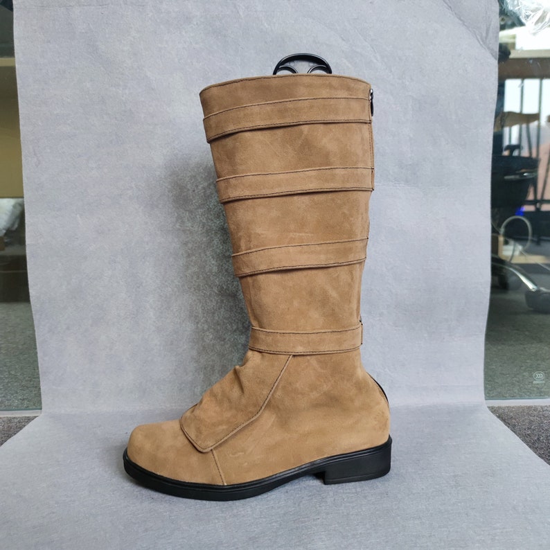 K&ouml;nnte beinhalten: Ein Paar braune, kniehohe Stiefel aus Wildleder mit mehreren Riemen und Schnallen. Die Stiefel haben eine schwarze Gummisohle.