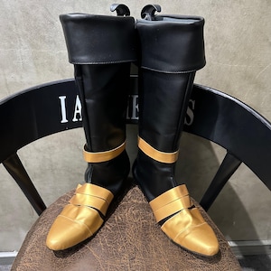 Puede incluir: Un par de botas negras y doradas hasta la rodilla. Las botas tienen una caña negra con una banda dorada y un pie dorado. La parte superior de la bota está doblada. Las botas están sobre una silla de cuero marrón.